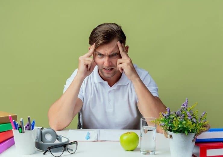 angry-young-handsome-male-student-sitting-desk-with-school-tools-putting-hands-forehead-isolated-olive-green_141793-63306